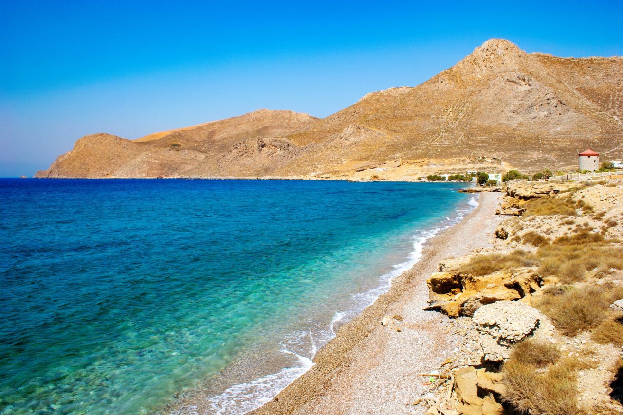 Crystal-clear blue water and a pebbled shoreline at Eristos Beach on Tilos Island, with dry rocky hills and a windmill in the background.
