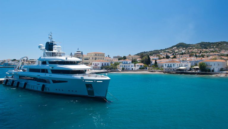 Luxury superyacht docked in the turquoise waters of Spetses, Greece, with a charming village and hills in the background.