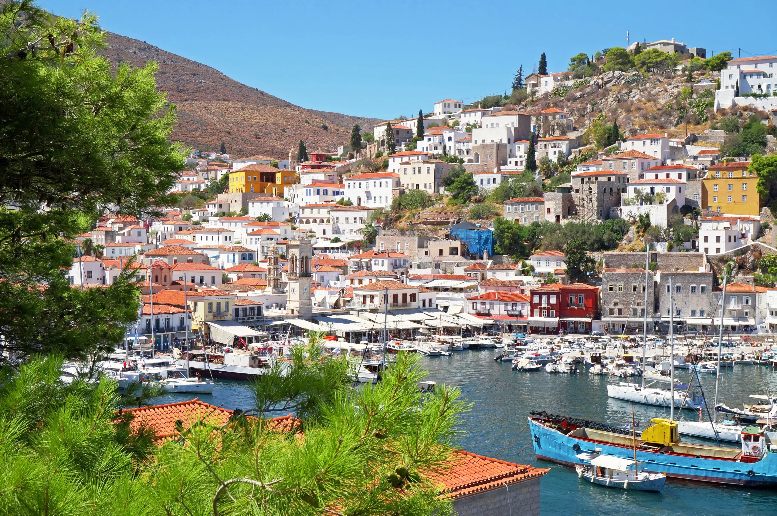Hydra harbor view with yachts, fishing boats, and colorful neoclassical buildings climbing the hillside.
