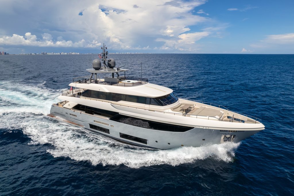 Modern luxury motor yacht cruising through open waters on a sunny day, with a coastal city skyline in the background.