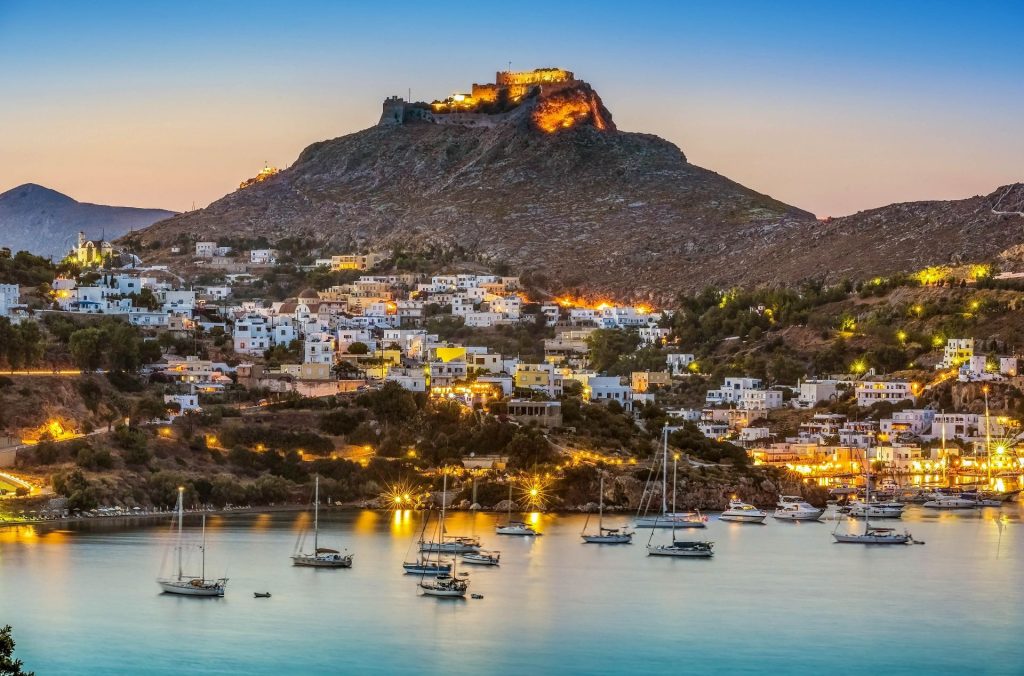 Evening view of Leros Island, Greece, with boats in the harbor and a hilltop fortress lit above the town.