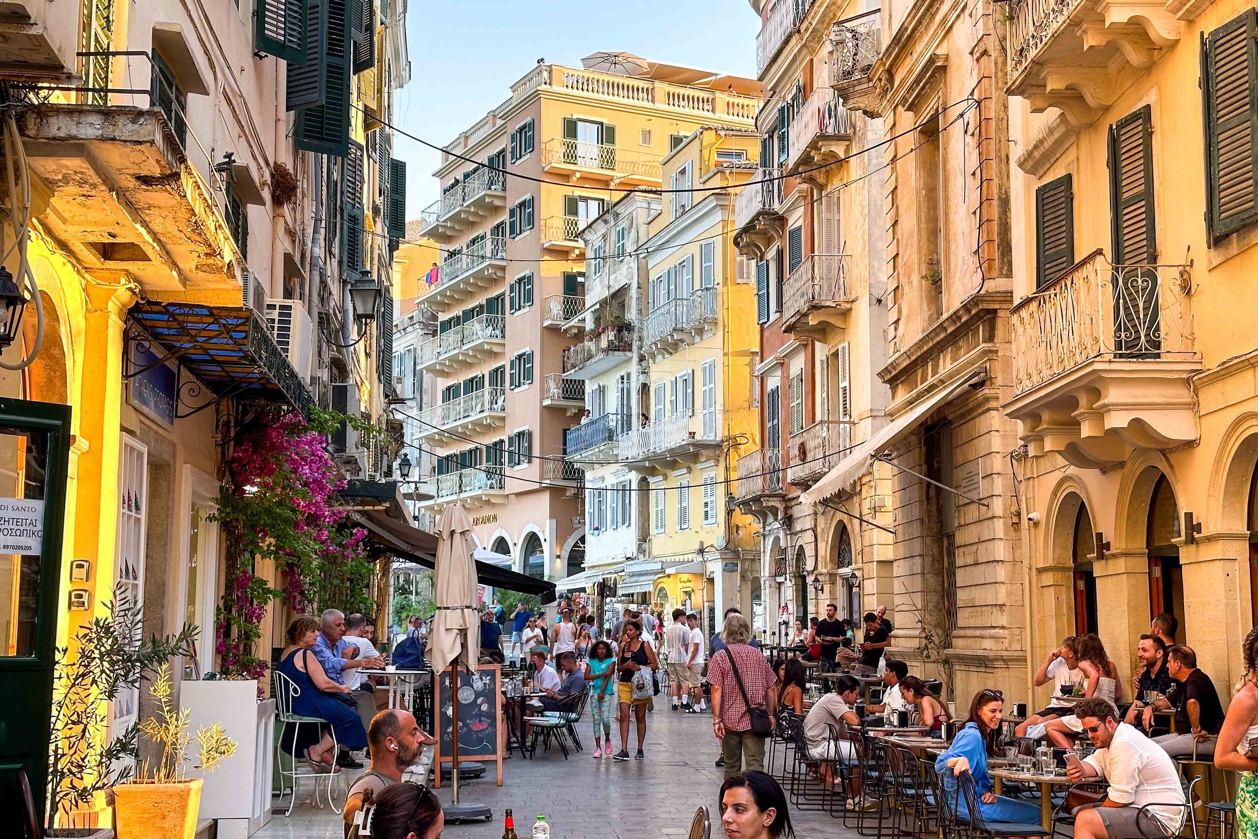People dining and walking along a lively pedestrian street surrounded by colorful Venetian-style buildings in Corfu&rsquo;s Old Town.
