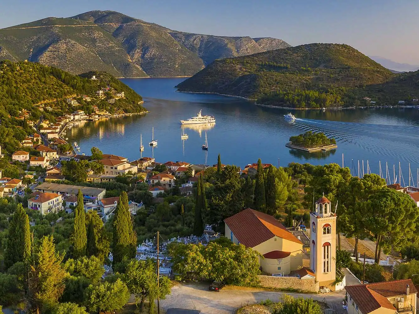 Aerial view of Vathi Port in Ithaca, Greece, with a bell tower, sailing yachts, and a white superyacht anchored in a calm bay surrounded by green hills.
