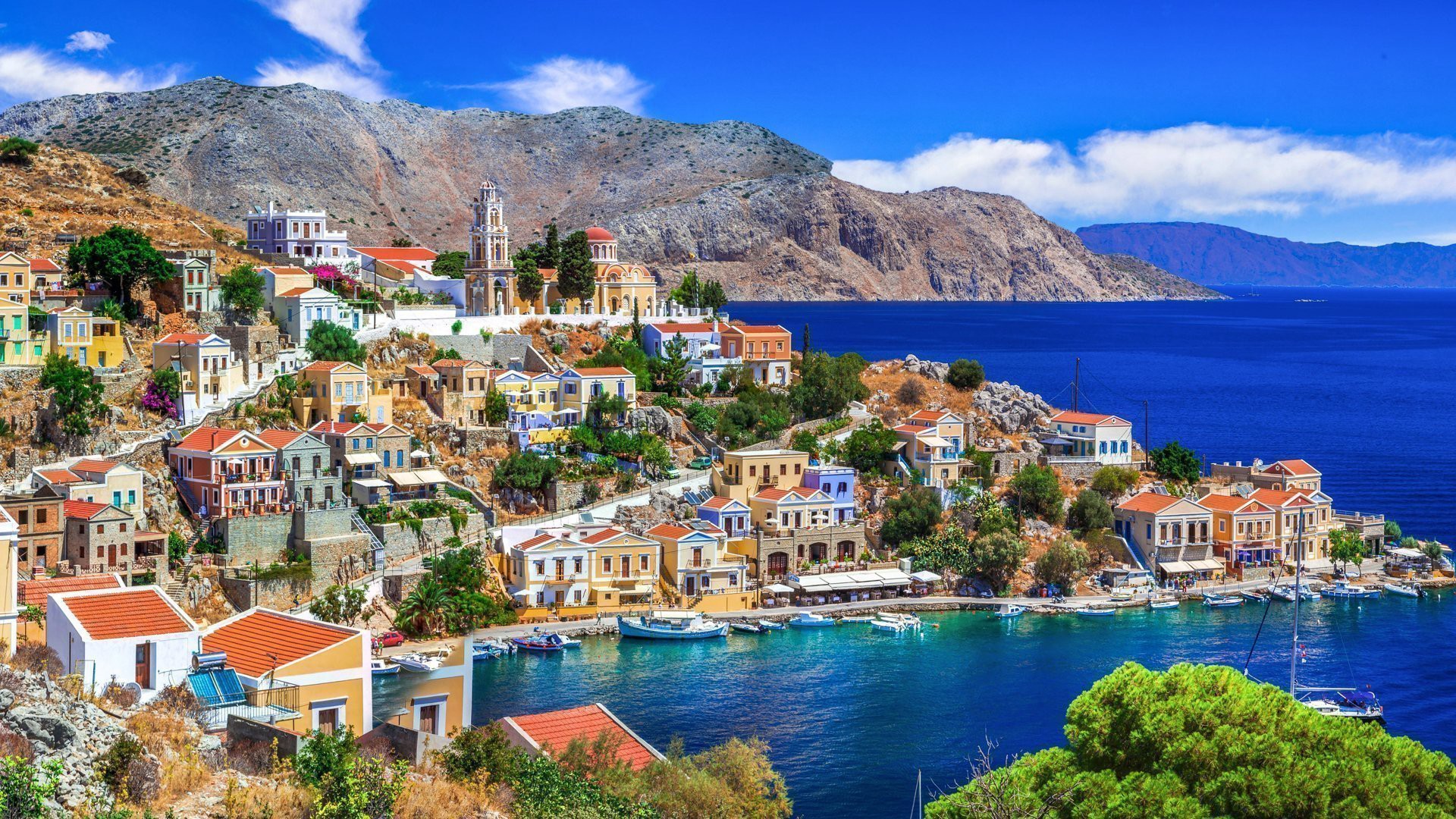 Colorful neoclassical houses on the hillside above a vibrant harbor in Symi Island, Greece, with deep blue sea and rugged mountains in the background.