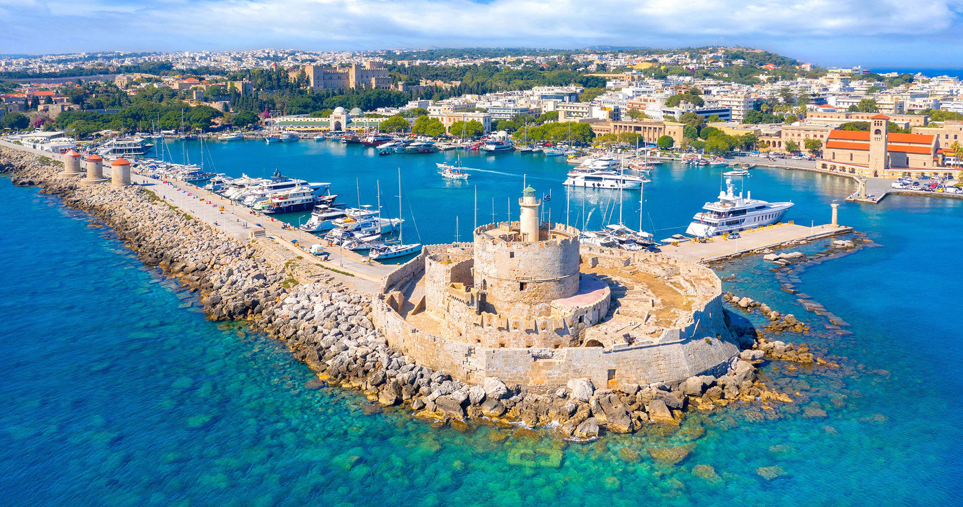 Aerial view of Rhodes Old Port with the medieval fort of Saint Nicholas, windmills, luxury yachts, and the city of Rhodes in the background.
