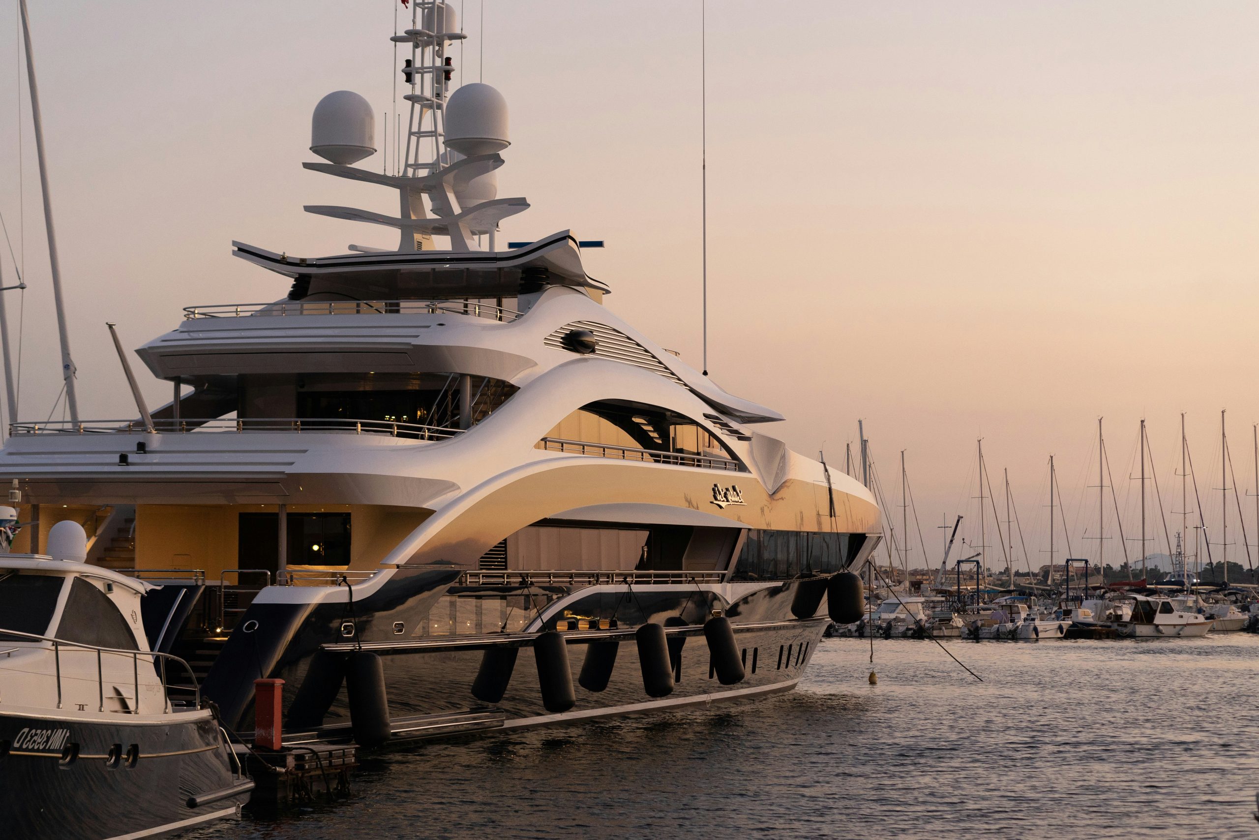 Luxury superyacht docked at a marina during sunset, with elegant curved design and surrounding sailboats in the background.