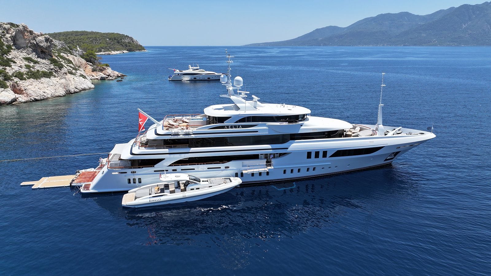 Superyacht AELIA anchored near a rocky coastline, with a Pirelli tender beside it and mountainous landscape in the background.
